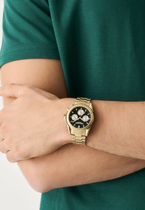 Asmuo, dėvintis auksinį rankinį laikrodį su juodu ir baltu ciferblatu, sukryžiuoja rankas ant žalia trumpomis rankovėmis marškinėlių.