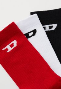 Trois paires de chaussettes mi-mollet en rouge, blanc et noir, chacune ornée d'un logo. Les chaussettes présentent un design tricoté texturé.