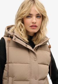 Steppweste in hellbeige mit Kapuze, ausgestattet mit einer glatten Textur, Knopfverschluss und Kordelzugakzenten. Über einem schwarzen Pullover getragen.