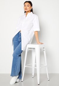 Camicia bianca oversize con bottoni e maniche corte, abbinata a jeans a zampa d'elefante blu chiaro e sneakers bianche, appoggiata a uno sgabello bianco.
