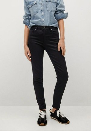 Person iført stramme sorte bukser, sort-hvide sneakers og en lyseblå denimskjorte med knapper og opkrogede ærmer.