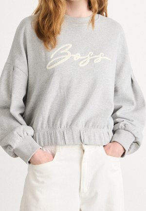Personne portant un sweat-shirt gris clair avec le texte « Boss » en écriture blanche, associé à un pantalon taille haute blanc cassé, mains dans les poches.