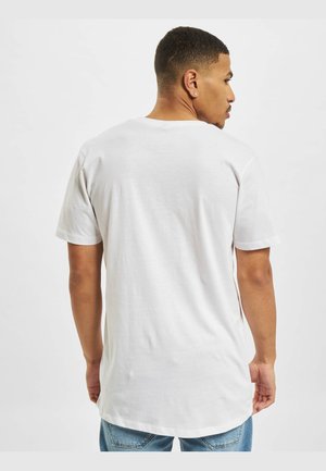 Muž s krátkymi vlasmi, oblečený v jednofarebnom bielom tričku s krátkymi rukávmi a modrých džínsoch, zobrazený zozadu na jednofarebnom pozadí.