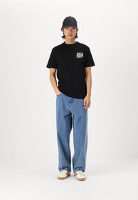 Schwarzes T-Shirt mit kurzen Ärmeln und grafischer Brusttasche, kombiniert mit lockeren blauen Jeans und weißen Schuhen mit roten Akzenten.