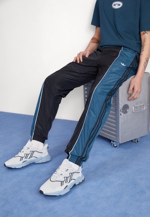 Osoba nosząca białe trampki, czarne i niebieskie spodnie dresowe z paskami oraz niebieską koszulkę, siedząca na metalowej skrzyni na kółkach.