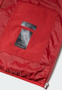 Intérieur d'une veste rouge montrant une poche cousue avec des étiquettes noires d'entretien et de performance, et une fermeture éclair partiellement visible le long du bord.