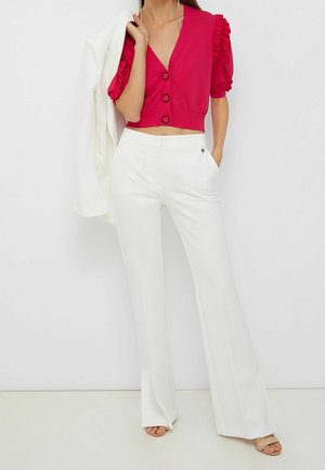 Vrouw die witte wijde broek met hoge taille draagt, een rode cropped blouse met geruite mouwen, en een wit jasje over haar schouder draagt.