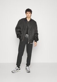 Zwarte bomberjack met een rits aan de voorkant, gecombineerd met zwarte joggingsbroeken met een wit logo, en metallic sneakers met een reflecterende afwerking.