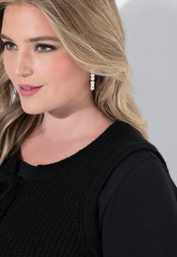 Svart stickad väst med rundad halsringning, bärs över en svart långärmad tröja. Utrustad med pärlhängörhängen och vågigt blont hår.