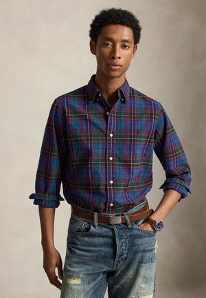 Jeune homme portant une chemise à carreaux bleue et rouge à boutons avec manches retroussées, jean déchiré, ceinture marron et montre-bracelet bleue.