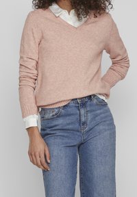 Ljusrosa V-ringad tröja i mjukt tyg, med ribbade mudd och nederkant, lagd över en vit skjorta med krage, bärs tillsammans med blå jeans.