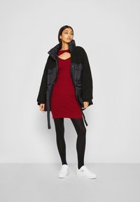 Giacca imbottita nera con texture in pelliccia sintetica sopra un vestito rosso a coste con una scollatura. Collant neri e sneakers bianche completano l'outfit.