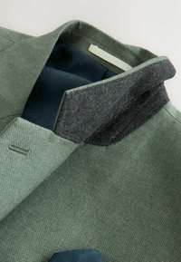 Colletto della giacca verde testurizzata ripiegato verso l'alto, che mostra la fodera interna grigio scuro e l'interno liscio della giacca blu navy con motivo.