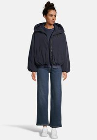 Doudoune oversize bleu marine avec capuche, fermeture éclair, poches latérales, ourlet côtelé et poignets élastiques, associée à un jean large et des baskets blanches.