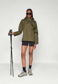 Πράσινο αδιάβροχο μπουφάν με φερμουάρ και κουκούλα, συνδυασμένο με μαύρο σορτς, γκρι παπούτσια πεζοπορίας και μπαστούνια πεζοπορίας, σε απλό φόντο.