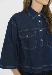 Ženska oblečena v temno modro skrajšano jeans jakno z širokimi rokavi, z gumbom na sprednji strani in dvema žepoma na prsih s kovinskimi gumbi.