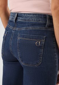 Tmavě modré rifle s vysokým pasem, hladkým povrchem, se dvěma zadními kapsami a malým kovovým logem na pravé kapse.