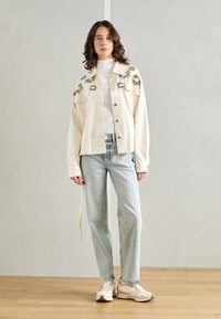 Kremfarget jakke med broderte blomsterdetaljer, knappestolpe, brukt over en hvit genser med høy hals, sammen med lyseblå jeans og hvite joggesko.