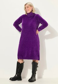 Abito a collo alto in maglia viola con maniche lunghe e tasche laterali. Indossato con stivali neri in pelle al ginocchio con suole spesse e cinturini.
