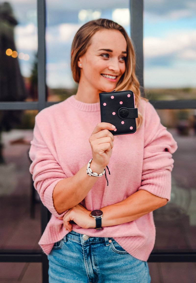 Black wallet with pink polka dots, featuring a circular clasp, held in a hand. Soft pink knit sweater and light blue jeans visible.