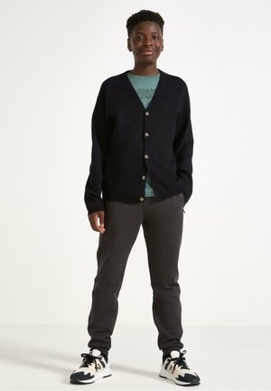 Mørkeblå cardigan med V-hals, der har fem lysere knapper. Sammen med en grøn T-shirt og grå sweatpants, afsluttet med sorte og hvide sneakers.