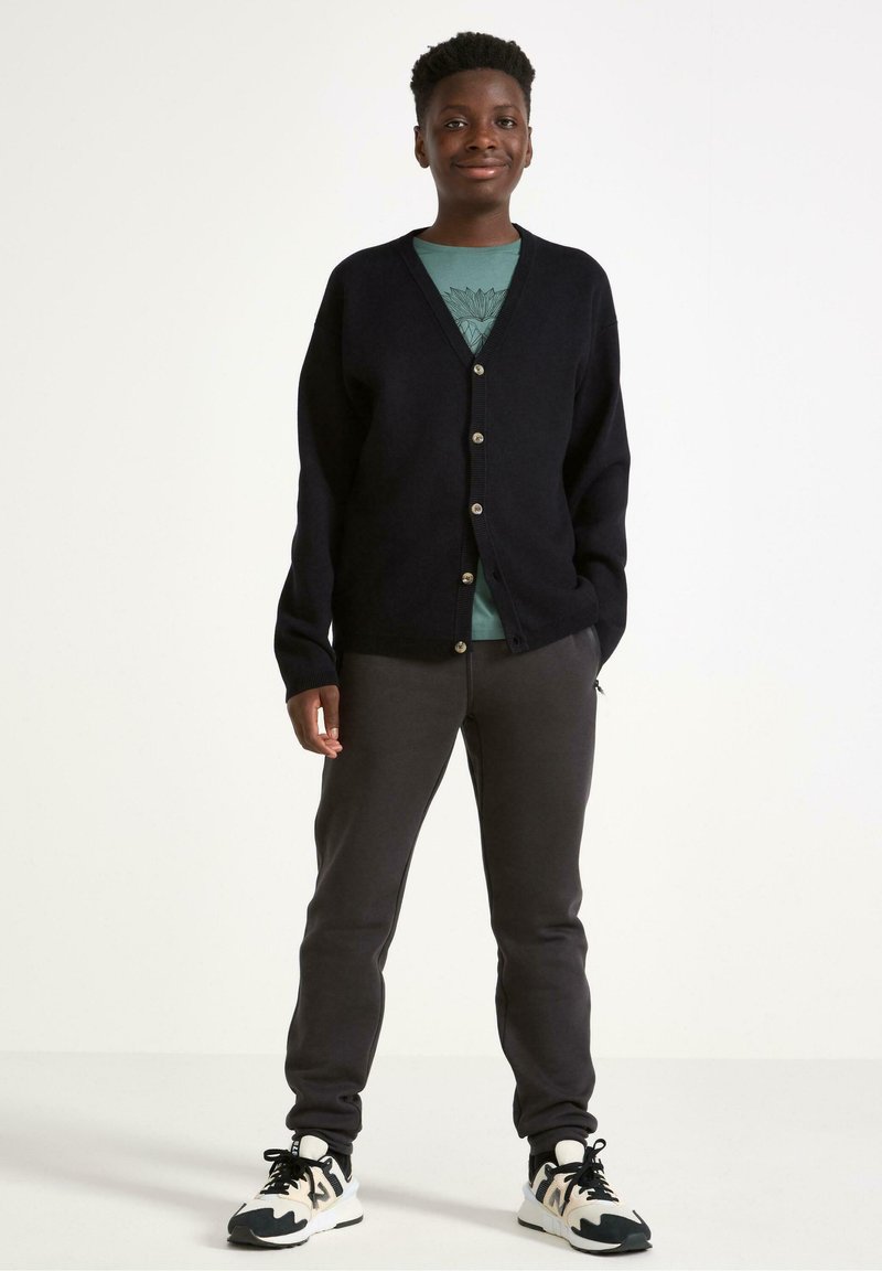 Mørkeblå cardigan med V-hals, der har fem lysere knapper. Sammen med en grøn T-shirt og grå sweatpants, afsluttet med sorte og hvide sneakers.