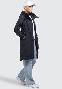 Marineblå vandtæt parka med snoretræk i taljen, lynlåslommer og høj krave. Bæres med lysblå brede bukser og hvide sneakers.