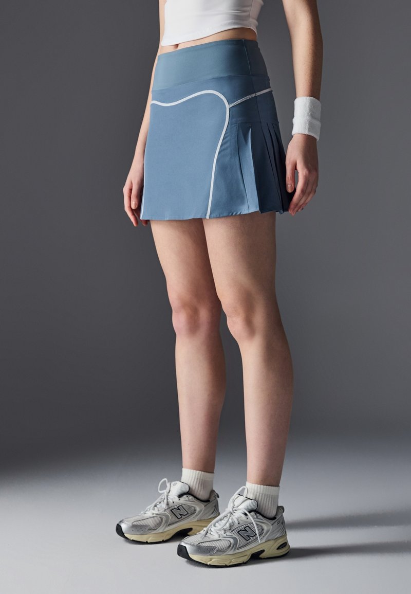 Femme portant une jupe de sport plissée bleue, un bracelet blanc, des chaussettes blanches et des baskets New Balance grises, debout sur un sol gris face à un fond sombre.