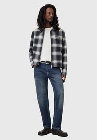 Harsohi plaid zip-up takki harmaassa ja valkoisessa, yhdistetty valkoisen paidan ja sinisten farkkujen kanssa. Musta vyö ja tummat kengät täydentävät asua.