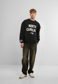 Svart hettegenser med teksten "NORTH CAROLINA" i hvit skrift, kombinert med mørke, løse jeans og hvite joggesko med lys brun såle.