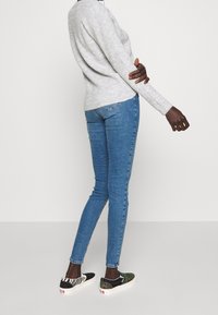Persona de pie de lado, vistiendo un suéter gris claro, jeans ajustados azules y zapatillas de cuadros desparejadas con patrones de cebra y camuflaje.