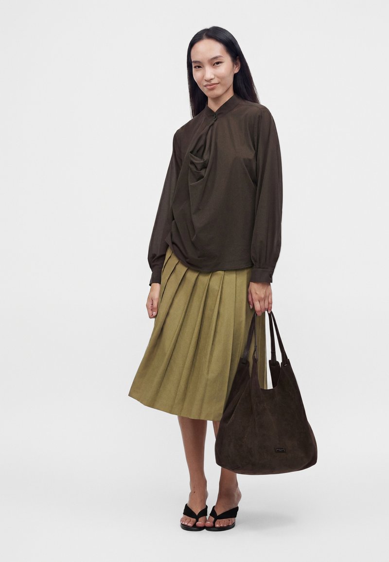 Femme portant un chemisier à manches longues marron foncé et une jupe plissée olive, tenant un grand sac fourre-tout marron foncé et portant des sandales noires.