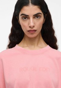 Ruzna pulover s okroglim izrezom, ki vsebuje besedilo "ROUGE EDIT" v subtilni, rahlo dvignjeni teksturi. Mehak material z udobnim krojem.