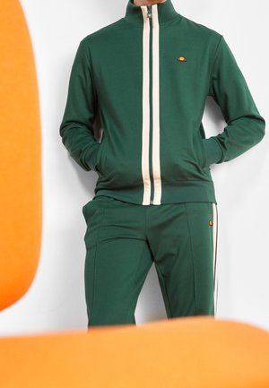 Person trägt einen dunkelgrünen Trainingsanzug mit weißen vertikalen Streifen und kleinen orangefarbenen Logos, die Hände in den Jackentaschen, steht vor einem weißen Hintergrund.