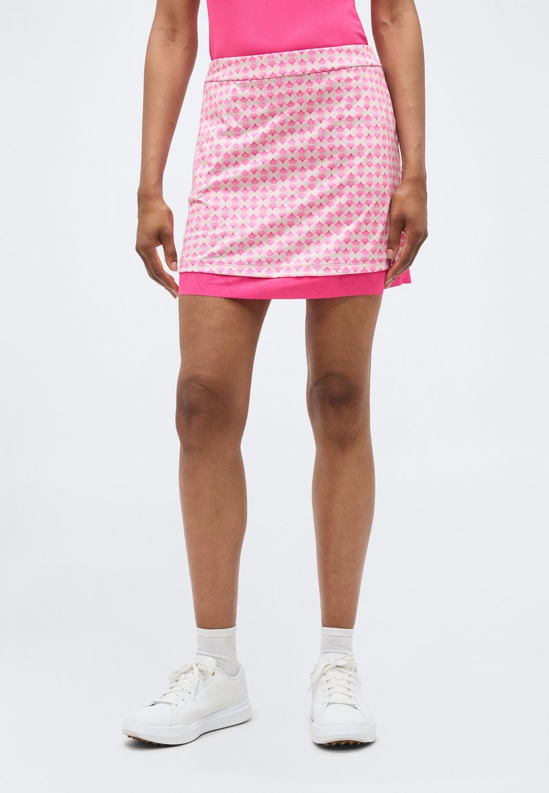 Vrouw die een roze mini-rok met patroon draagt over een effen roze rok, witte enkelsokken en witte sneakers, staand tegen een effen achtergrond.