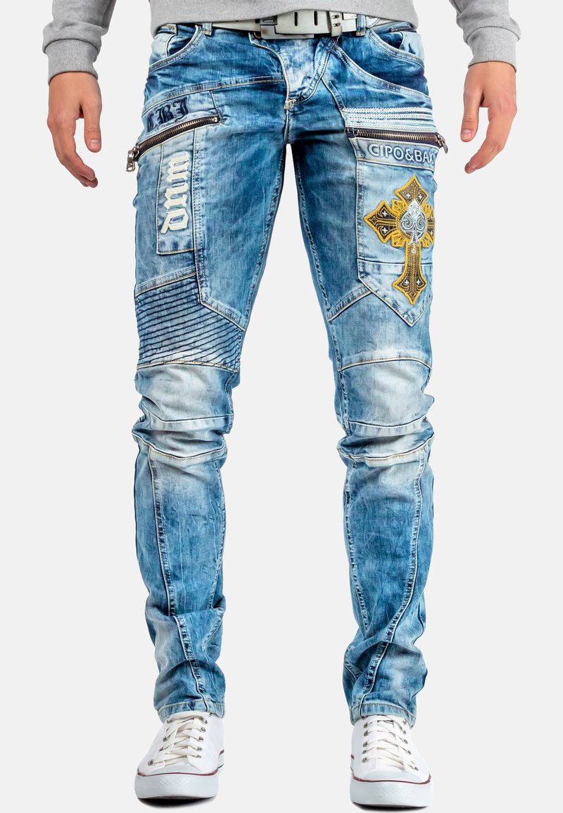 Jeans in denim blu chiaro con taglio slim, design a croce ricamato, numerose tasche con zip e pannelli testurizzati sulle gambe.