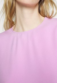 Blusa rosa a collo alto con texture liscia e scollatura cucita. Il tessuto presenta una leggera lucentezza, creando un aspetto raffinato.