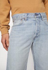 Man wearing light blue high-waisted denim jeans and a brown long-sleeve shirt, hand relaxed by side.