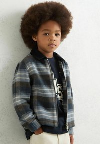 Dryžuota striukė su užtrauktuku mėlynos, pilkos ir baltos spalvų atspalviuose, su juoda šonkauline apykakle, derinama su tamsiai mėlynu marškinėliu, dėvima vaiko.