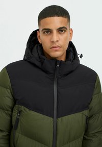 Giacca imbottita con sezione superiore nera e sezione inferiore verde, dotata di un alto colletto, chiusura con zip e tasche laterali.