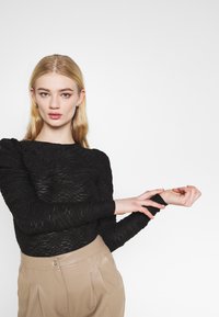 Blusa preta de mangas compridas com padrões texturizados em ondas; acompanhada por calças beiges claras de cintura alta, com detalhe de botão.