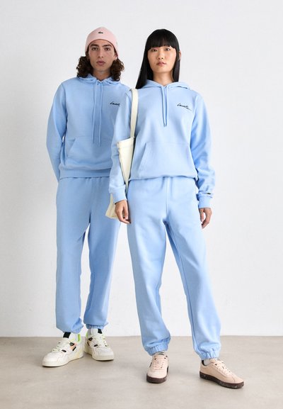 Sweatshirts à capuche et joggers assortis bleu clair, présentant des poches avant et des poignets côtelés. Associés à des baskets et un sac fourre-tout.