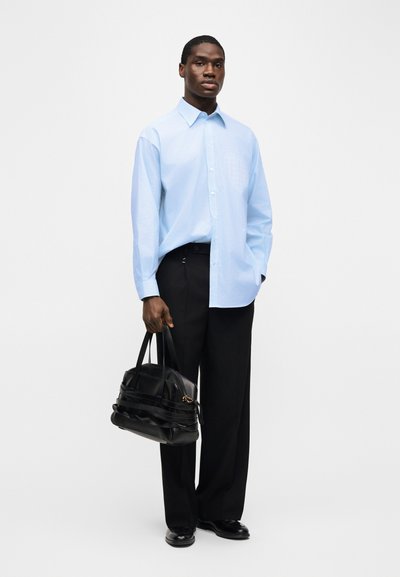Άνδρας φορώντας ανοιχτό μπλε πουκάμισο με κουμπιά και μαύρο παντελόνι, κρατώντας μαύρη δερμάτινη τσάντα, στέκεται μπροστά σε απλό λευκό φόντο.