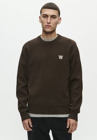 Brauner Pullover mit geripptem Ausschnitt und Bündchen, mit einem kleinen weißen Logo auf der Brust. Über einem grauen T-Shirt getragen.
