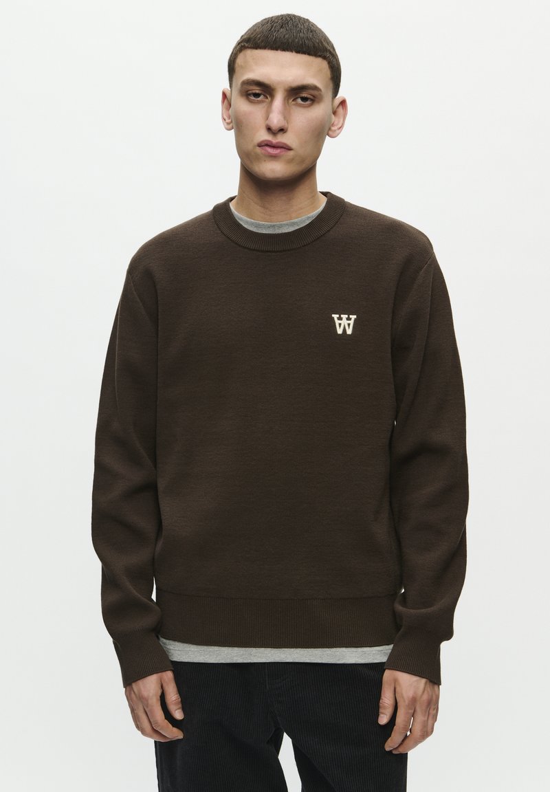 Brauner Pullover mit geripptem Ausschnitt und Bündchen, mit einem kleinen weißen Logo auf der Brust. Über einem grauen T-Shirt getragen.