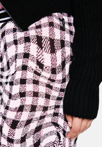 Hand auf der Hüfte, trägt einen schwarzen gerippten Ärmeln; karierte pinke und schwarze Rock mit einem kleinen, sichtbaren "Maddow Sports"-Label.