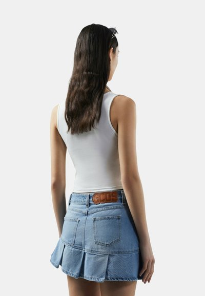 Vrouw met lang donker haar draagt een wit mouwloos topje en een geplooide blauwe denimrok met een GCDS-logo op de achterkant van de tailleband.