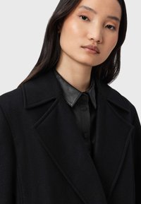 Cappotto nero in lana con grandi risvolti e tessuto strutturato, indossato sopra una camicia nera in pelle con colletto e bottoni a pressione.