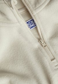 Felpa con cappuccio color crema in pile, con chiusura a zip, caratterizzata da un tessuto testurizzato, una zip liscia e un'etichetta blu all'interno del collo.