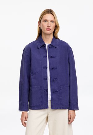 Femme aux longs cheveux blonds porte une veste ample bleu foncé avec de grandes poches avant, chemise blanche et pantalon beige clair, debout sur un fond blanc.